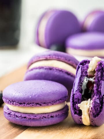 Blueberry macarons stacked on a wooden board with more macrons in the background.