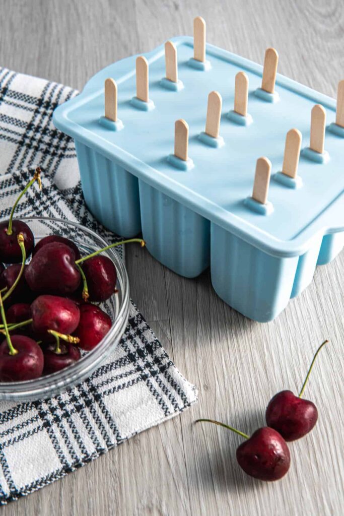 Cherry mango popsicles in a popsicle mold next to a bowl of cherries.