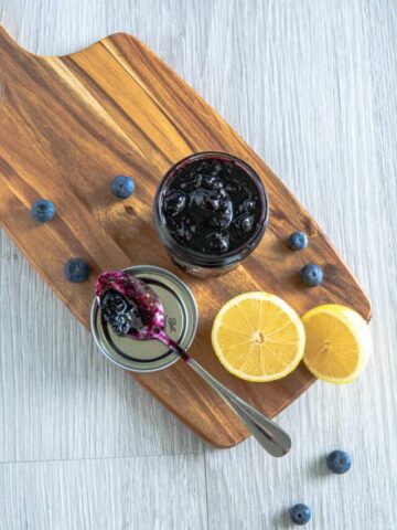 A jar of blueberry vanilla jam on a wooden serving board with lemons and a spoon.