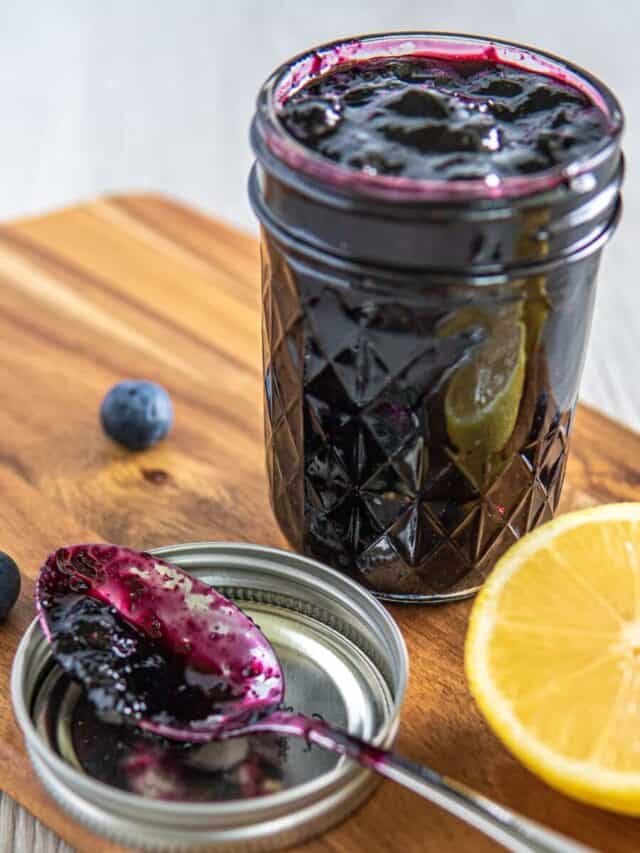 A jar of vanilla blueberry jam on a wooden serving board next to a spoon and half a lemon.