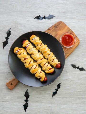 Halloween mummy hot dogs on a black plate with ketchup and bats.