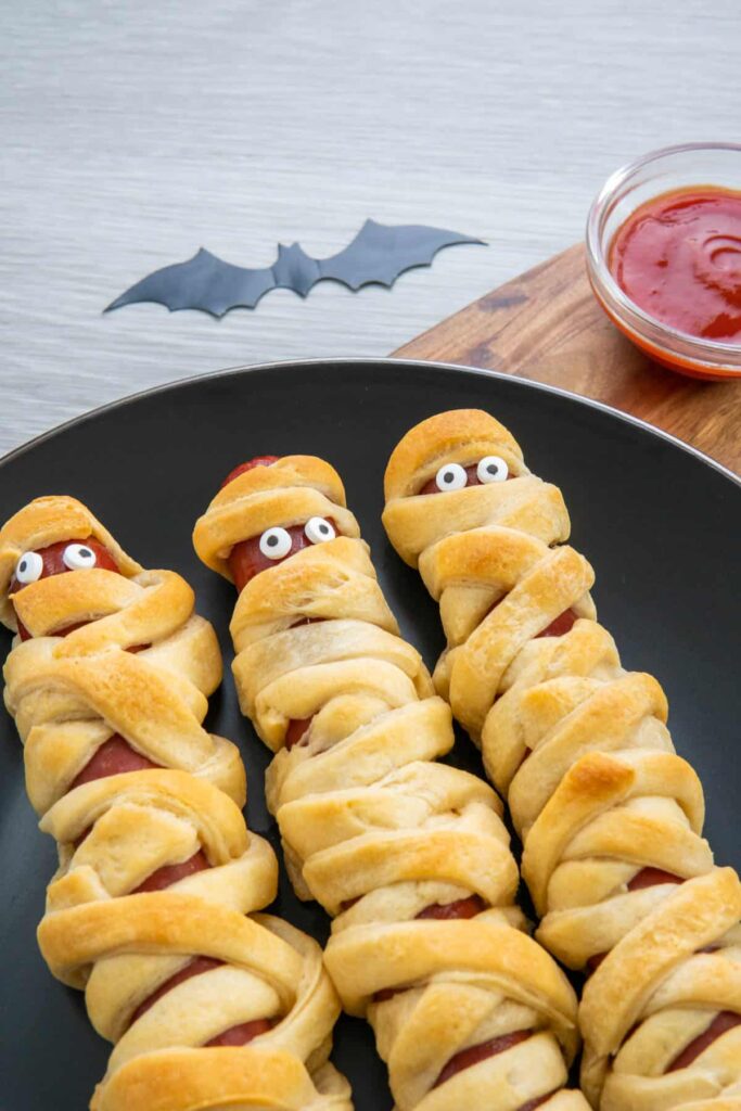 Halloween mummy hot dogs on a black plate with ketchup.