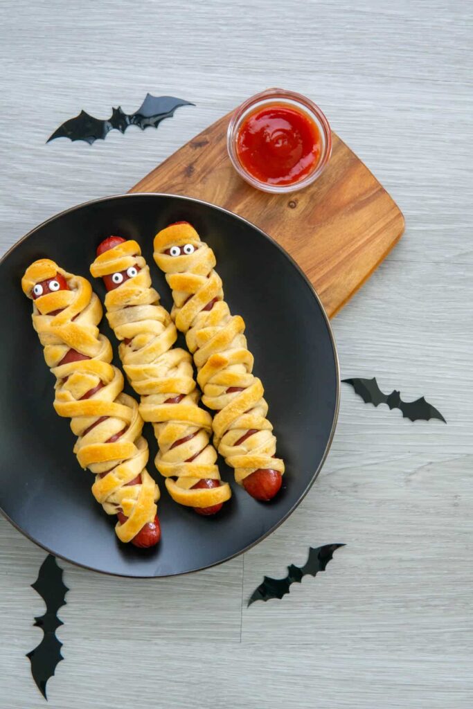 Mummy dogs on a black plate with bats scattered around and a bowl of ketchup.