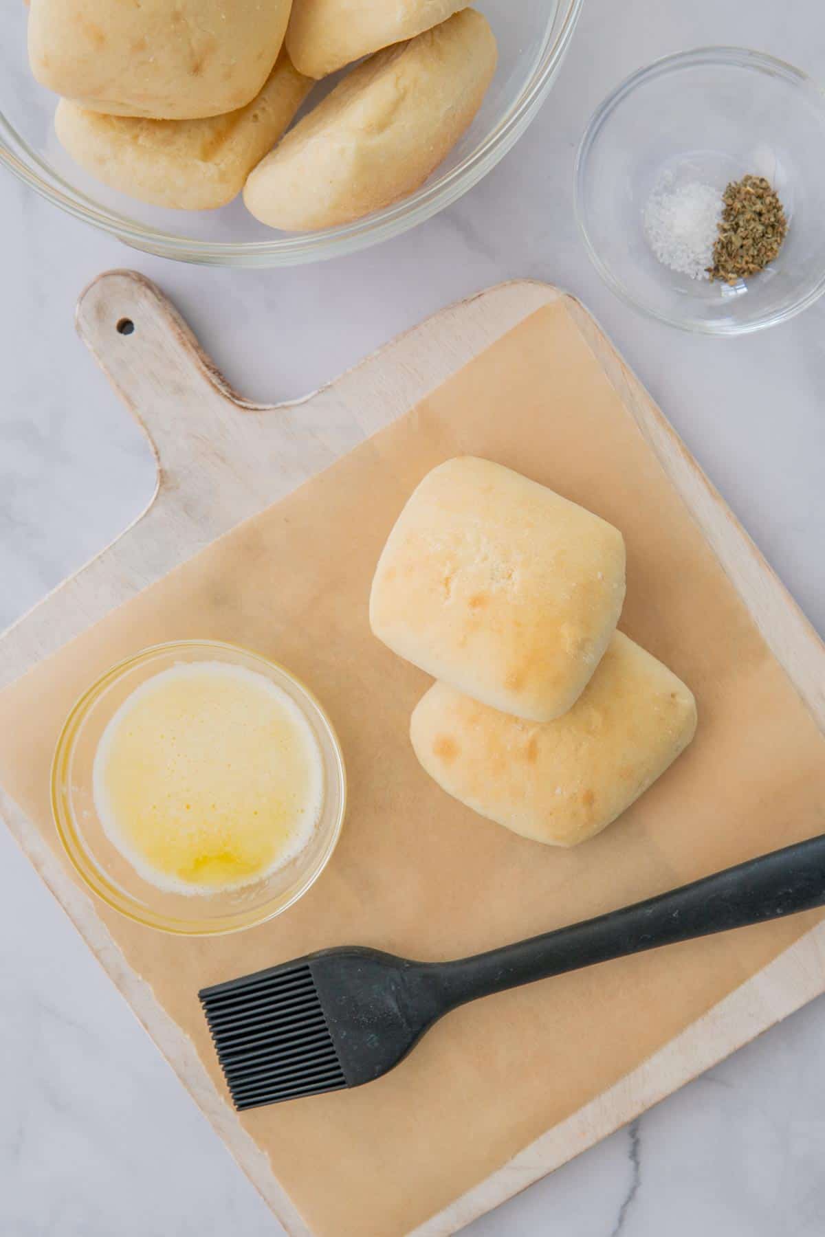 Dinner rolls on a wooden board next to a bowl of melted butter and a pastry brush.