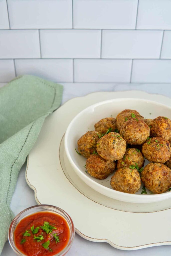 Air fryer turkey meatballs in a white bowl with marinara sauce and a green napkin in front of a white subway tile backdrop.