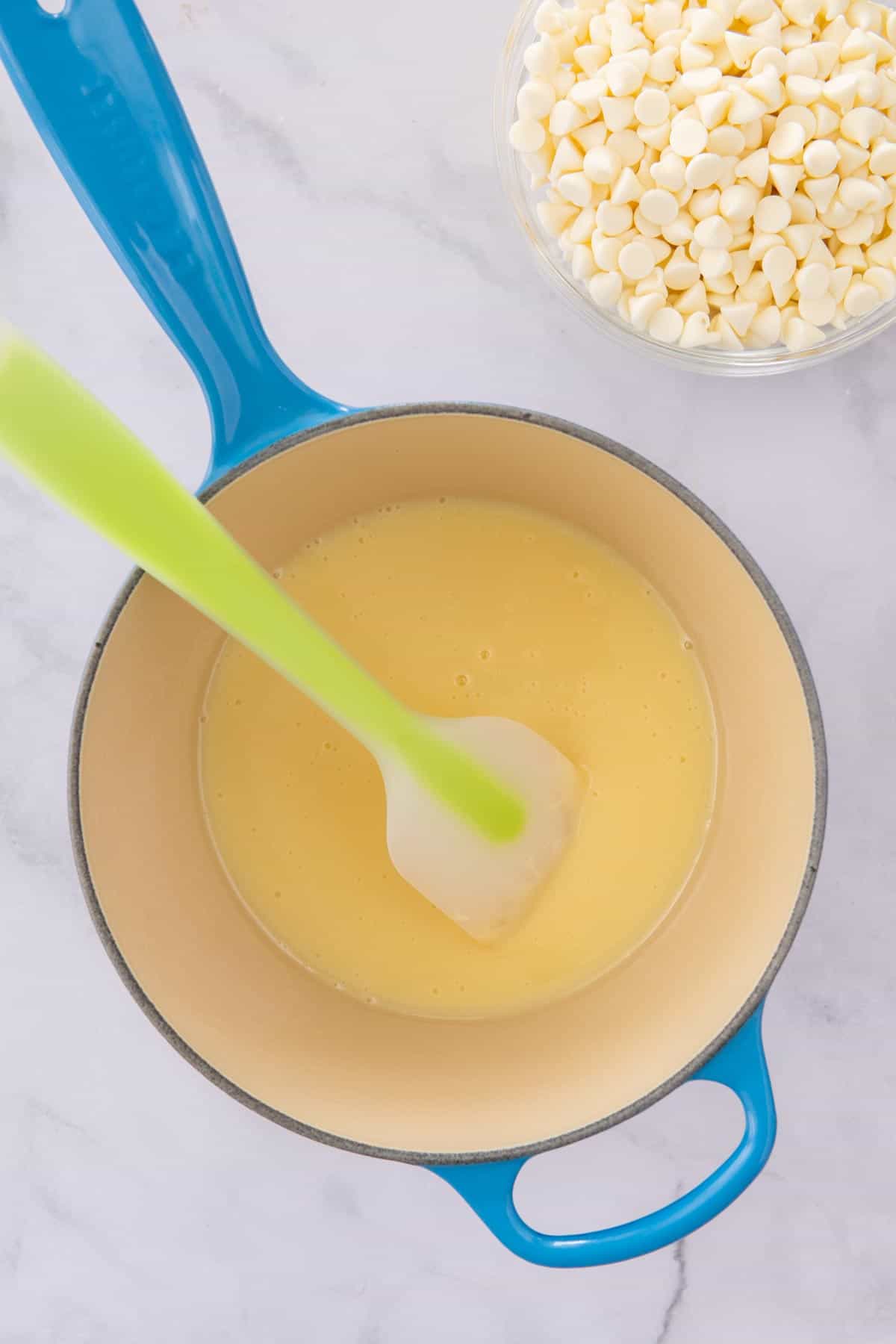 Condensed milk in a blue Le Creuset saucepan next to a bowl of white chocolate chips.