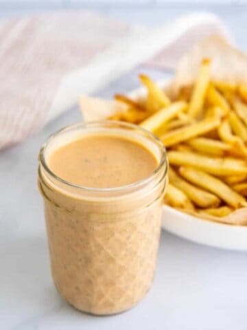 A close-up of a jar of BBQ ranch dressing with french fries in the background.