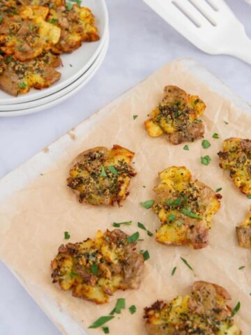 Air fryer smashed potatoes on parchment with a plate of potatoes in the background.