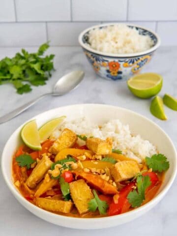A close up of Vegan Thai red curry with tofu in a white bowl with rice, lime, and cilantro.