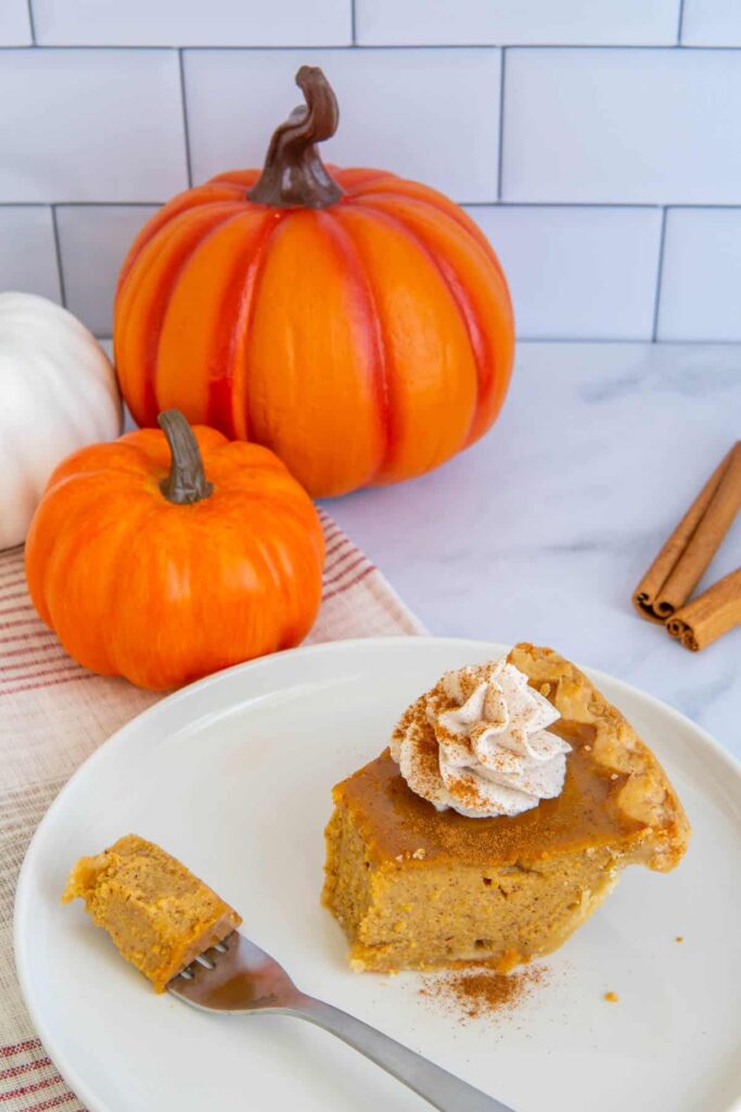 A slice of pumpkin pie with a bite taken out on a white plate with pumpkins in the background.