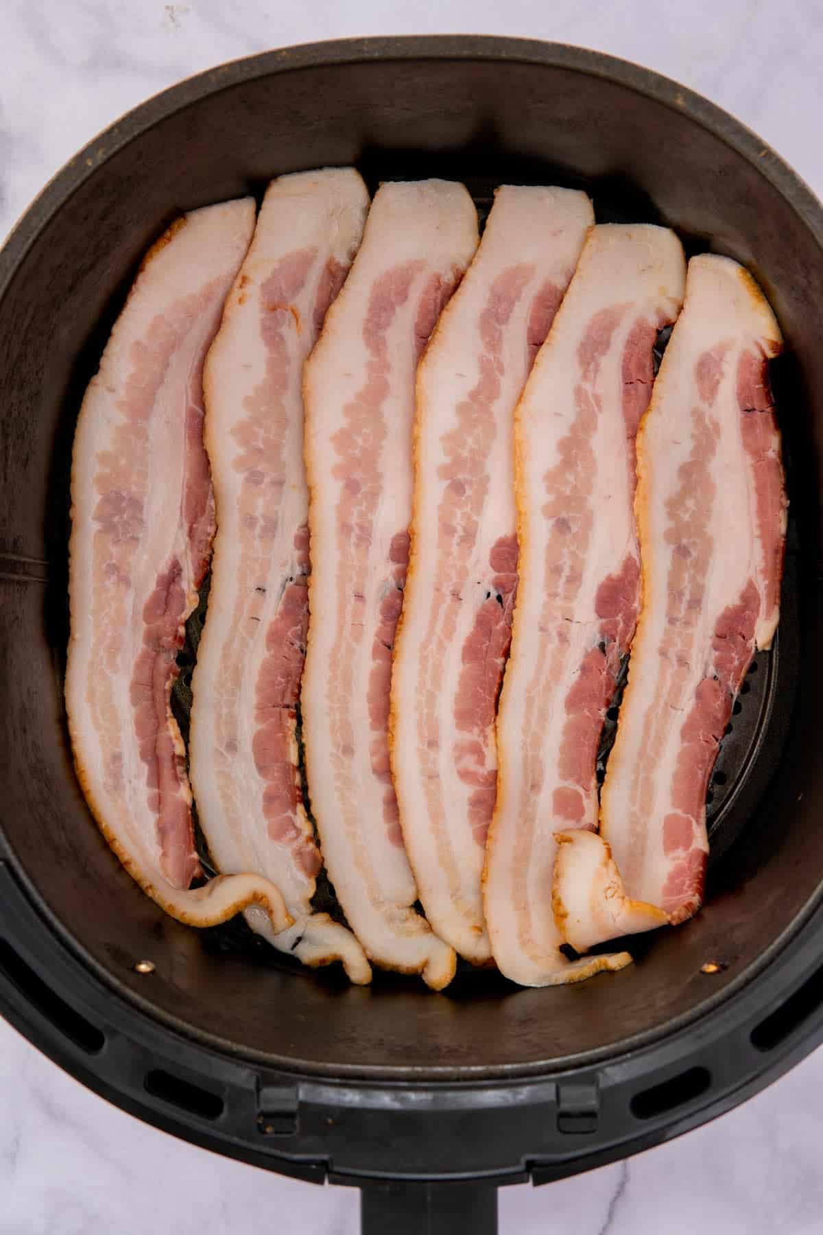 Uncooked strips of bacon in an air fryer basket.