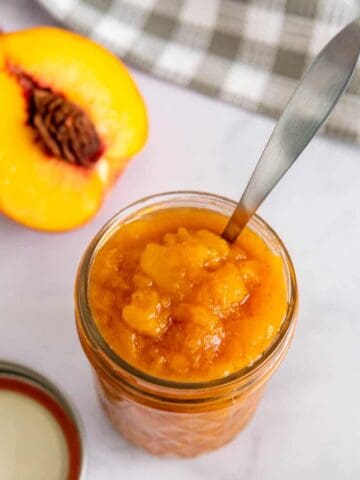 Peach jam in a glass jar with a spoon and fresh peaches.