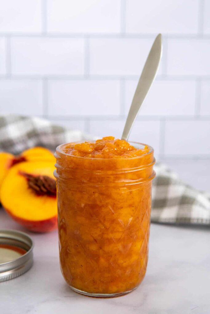 A jar of peach jam without pectin with fresh peaches and a napkin in the background.