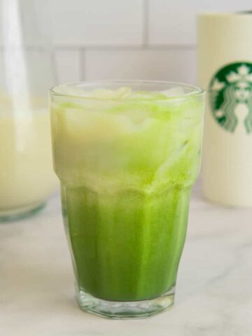Oat milk matcha latte in a glass with oat milk and a Starbucks cup in the background.