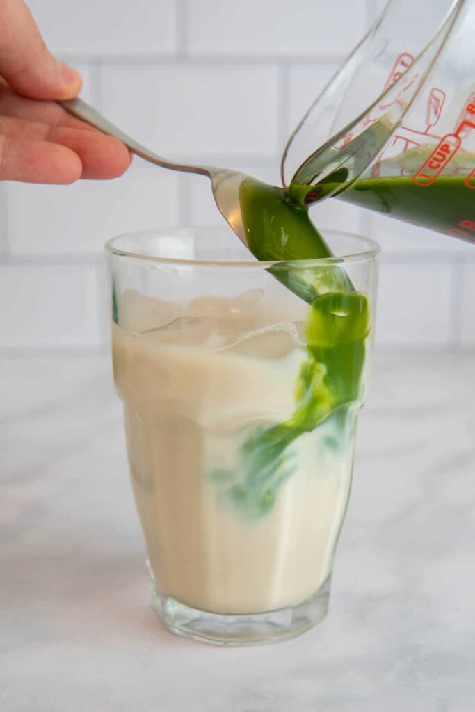 Pouring matcha over a spoon into a glass of oat milk and ice.