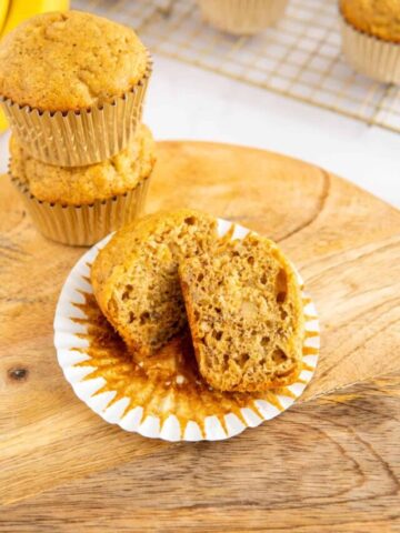 A banana bread muffin cut in half in front of a stack of two muffins.