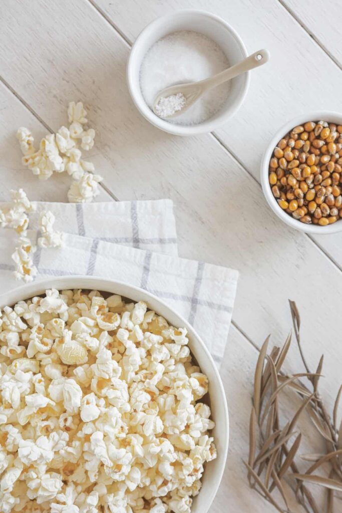 Popped popcorn in a bowl with popcorn kernels and a bowl of salt. 