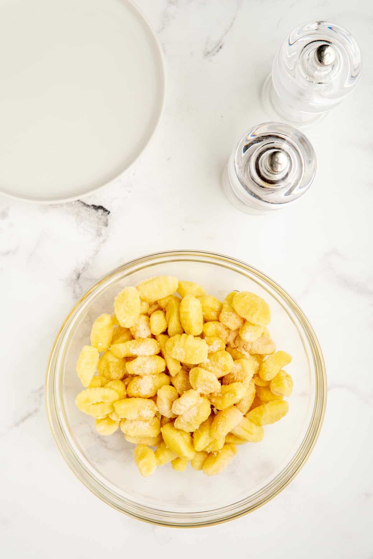 Uncooked gnocchi in a glass bowl with oil and seasonings sprinkled on top.