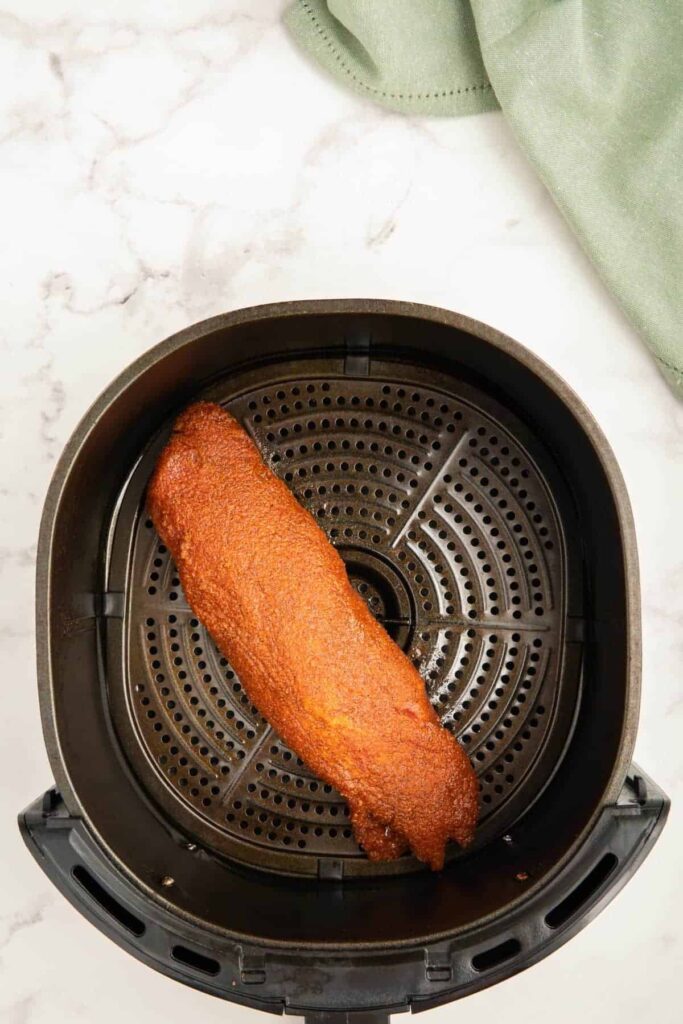 A raw pork tenderloin coated in oil and seasoning in an air fryer basket.