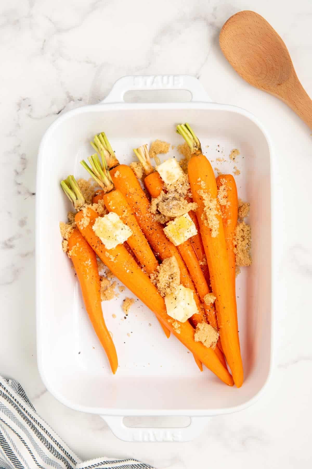 Raw carrots with brown sugar, honey, and butter in a white baking dish.
