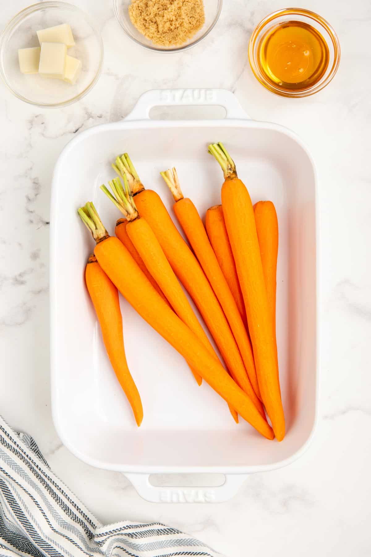 Raw carrots in a white baking dish with honey, brown sugar, and butter in glass bowls.