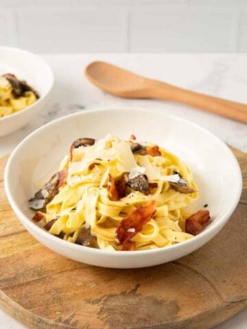 Bacon and mushroom carbonara in a white bowl on a wooden board with a wooden spoon in the background.