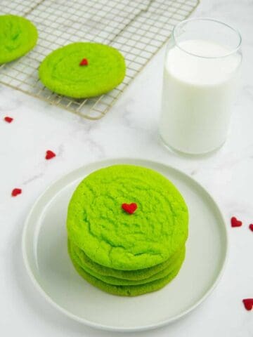 Grinch sugar cookies on a white plate with a glass of milk and more cookies in the background.