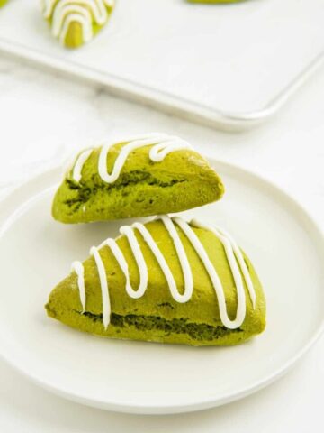 Two matcha scones with white chocolate drizzle stacked together on a white plate.