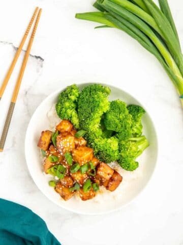 An overhead shot of sticky tofu in a white bowl with rice and broccoli.