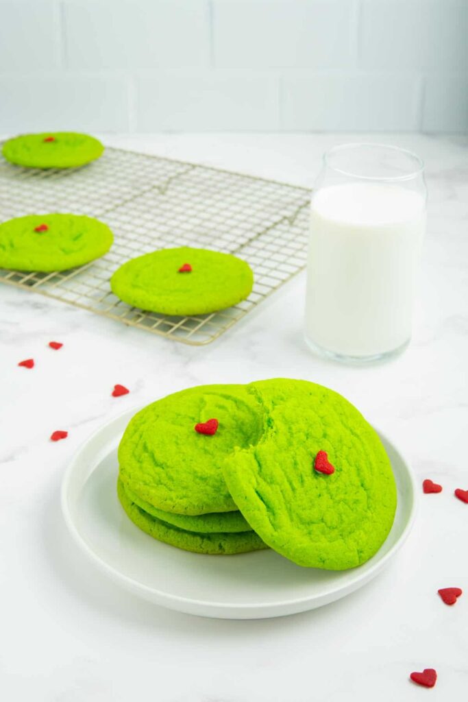A stack of three Grinch sugar cookies on a white plate with a fourth cookie with a bite taken out.