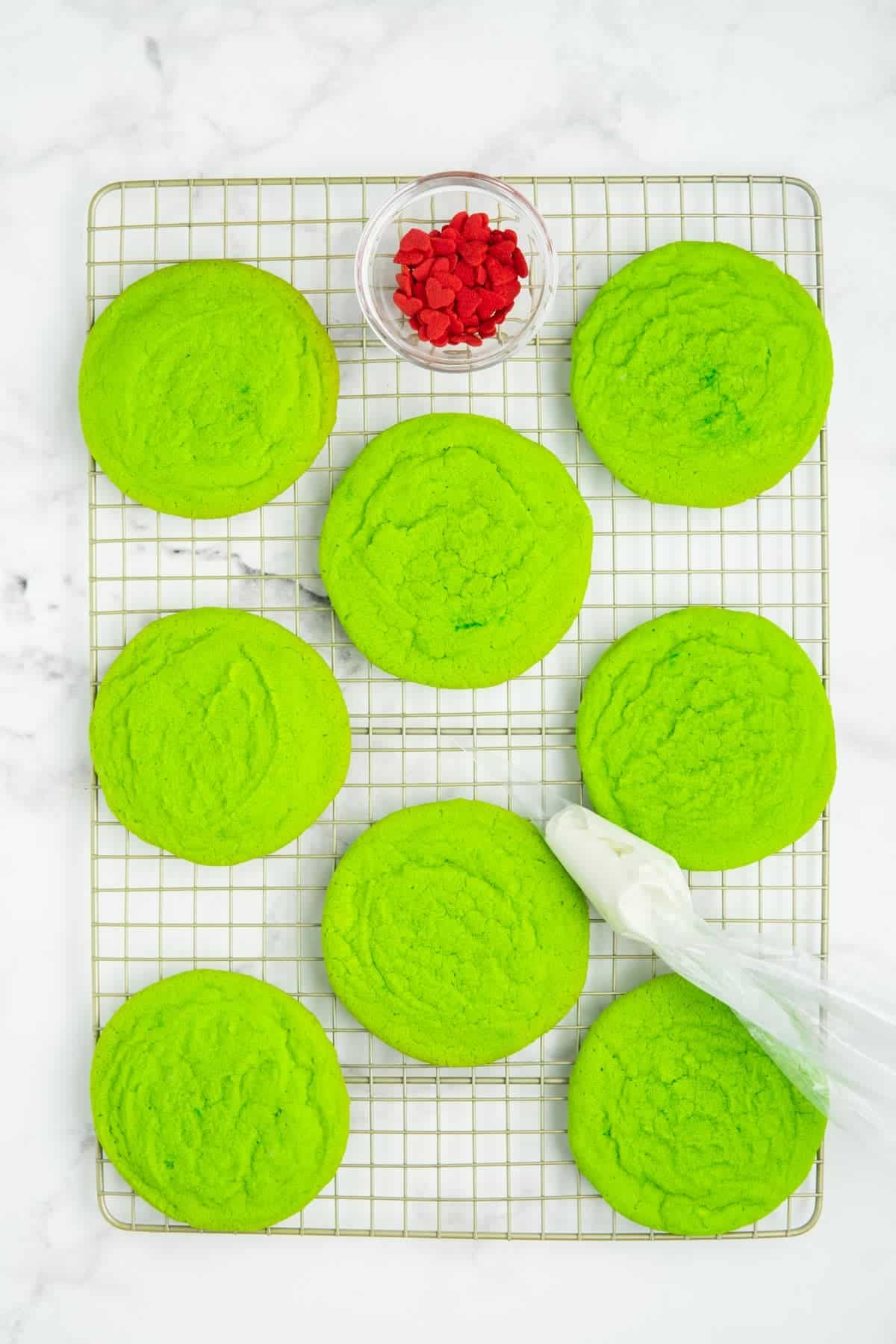 Grinch sugar cookies on a wire cooling rack with a piping bag of melted white chocolate.