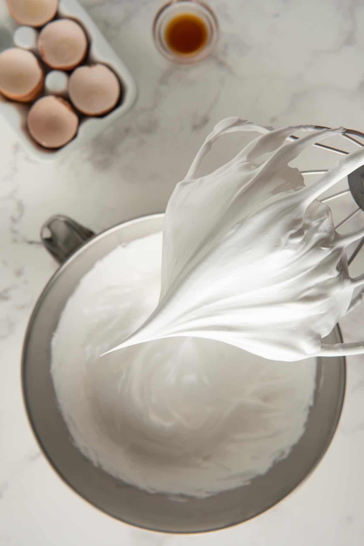 Meringue on a whisk at the stiff peaks stage above a mixing bowl.