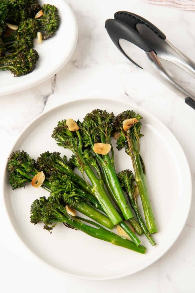 Roasted tenderstem broccoli and garlic on a round white plate next to tongs.