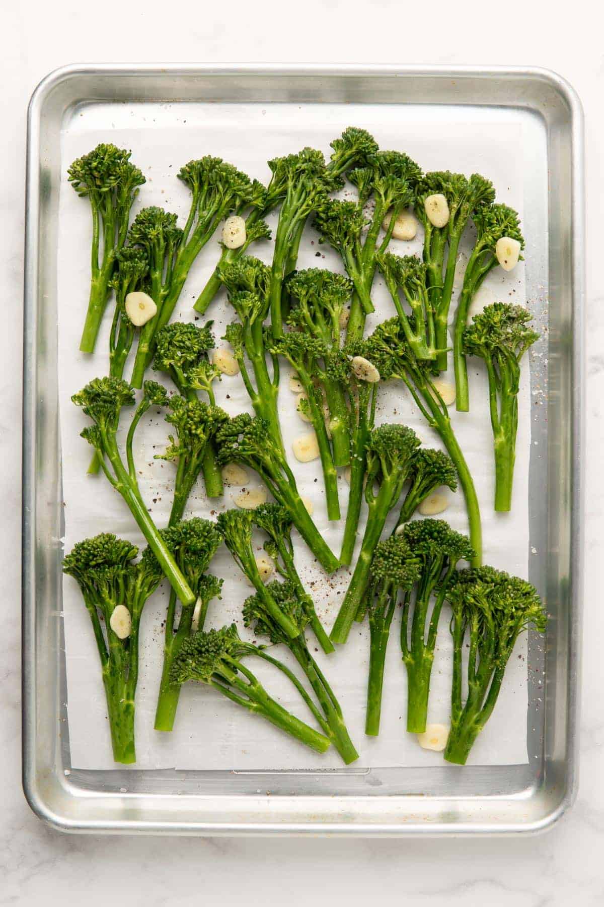 Uncooked tenderstem broccoli with garlic slices and seasoned with salt and pepper on an oven tray.