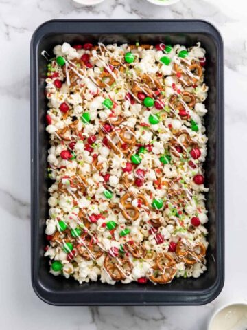 Christmas popcorn mix with pretzels and M&M candy drizzled with white chocolate and topped with sprinkles.