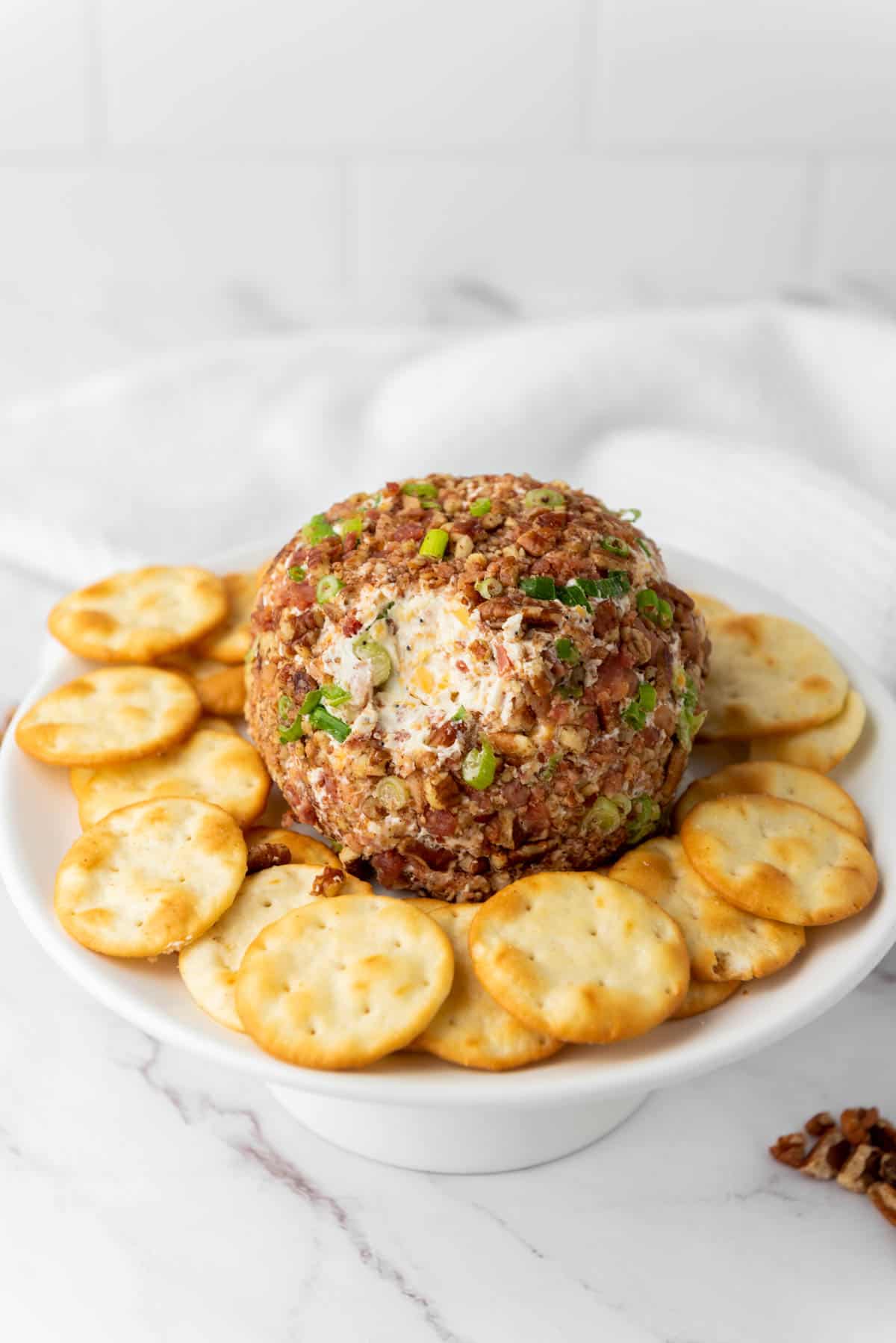 A pecan bacon cheese ball served with crackers on a white stand.