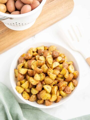 Air fryer diced potatoes in a white bowl with a colander full of uncooked potatoes in the background.