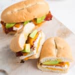 A chicken bacon sandwich sliced in half on a wooden board with another sandwich in the background.