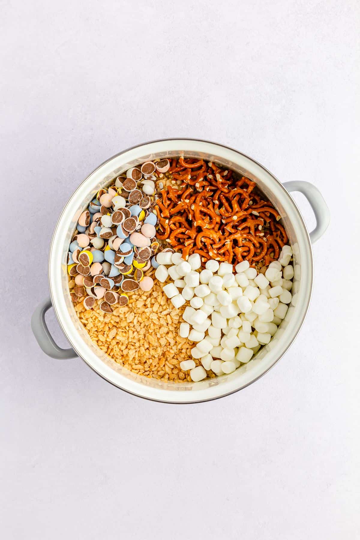 Adding rice krispie cereal, mini pretzels, mini eggs, and marshmallows to make rice krispie treats.