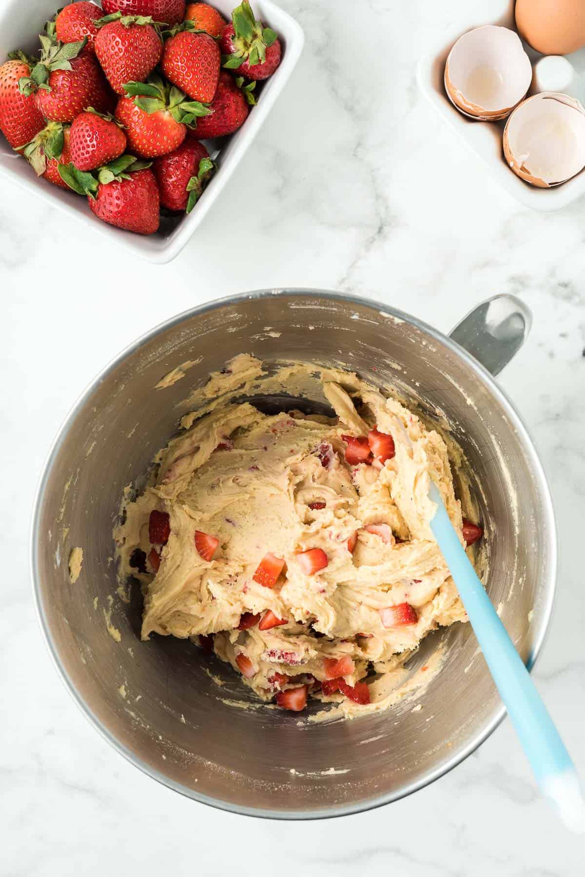 Adding fresh strawberries to cookie dough in a stainless mixing bowl.