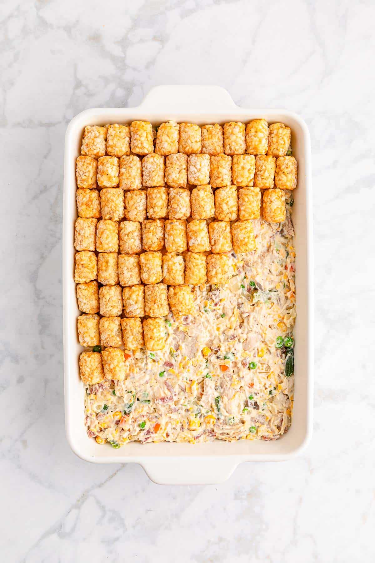 Topping chicken mixture with tater tots in a white baking dish.